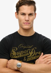 Sort bomulds T-shirt med logo i guldtryk, udstyret med korte ærmer. Et rundfacet ur med brun læderrem bæres om håndleddet.