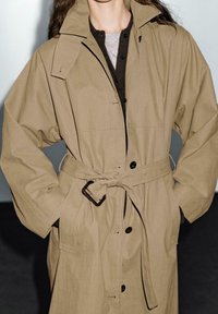 Manteau trench beige avec taille ceinturée, grand col et fermeture à boutons. Dispose de manches amples et de poches avant avec une texture lisse.