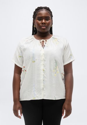 Femeie cu părul împletit poartă o bluză albă lejeră cu nasturi, cu broderie florală, și pantaloni negri, pe un fundal simplu, deschis la culoare.