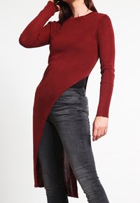 Burgunderfarbene Langarmbluse mit asymmetrischem Saum, gerippter Textur und figurbetonendem Schnitt, kombiniert mit dunklen Skinny-Jeans.