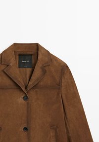 Giacca in camoscio marrone con colletto a revers, chiusura con un bottone e tasche laterali. Presenta dettagli di cuciture e un'etichetta all'interno.