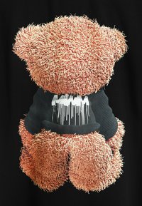 Diseño de oso de peluche con pelaje marrón texturizado y desaliñado; lleva una camiseta negra con un gráfico de pintura blanca goteando. Fondo negro.