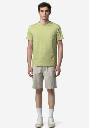 Giovane uomo in piedi che indossa una t-shirt verde chiaro con tasca, pantaloncini beige, calzini bianchi e scarpe da ginnastica bianche su uno sfondo bianco semplice.