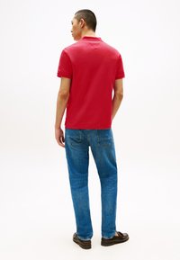 Κόκκινο πόλο πουκάμισο με κλασικό γιακά και κοντά μανίκια, συνδυασμένο με μπλε τζιν. Το σύνολο ολοκληρώνεται με σκουρόχρωμα παπούτσια.