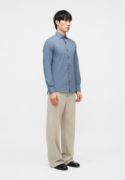 Άνδρας που στέκεται στραμμένος προς τα δεξιά, φορώντας στενή μπλε πουκάμισο με κουμπιά, μπεζ παντελόνι φαρδιάς γραμμής και μαύρα παπούτσια σε λευκό φόντο.