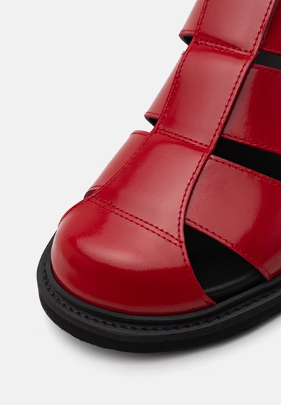 Punainen patenttikenkä, jossa on avonainen varvasosa, vaakasuuntaisia ja pystysuuntaisia hihnoja sekä musta pohja, jossa on ompeluyksityiskohtia.