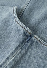 Tissu en denim bleu clair, présentant une texture lisse et des coutures visibles, avec un petit détail de fermeture éclair contrastant près de l'ourlet.