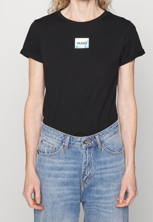 Osoba ubrana w czarny T-shirt z krótkim rękawem, na którym znajduje się mała naszywka z logo „HUGO” na środku, oraz w jasnoniebieskie jeansy z wysokim stanem.