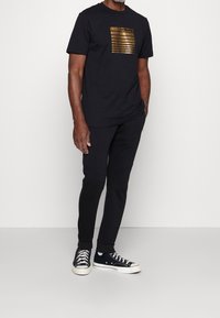 Black t-shirt with gold horizontal stripes and "MORATO" text, paired with black joggers and black sneakers featuring white soles.