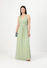 Világos zöld maxi ruha V-nyakkivágással, redőzött derékkal és könnyed szoknyával. A modell egy kis kék táskát tart. Simáva szövet, puha tapintással.