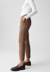 Pantaloni in suede marrone con tasche laterali inclinate, vestibilità slim e cuciture visibili, abbinati a mocassini neri e una maglietta bianca a maniche lunghe.