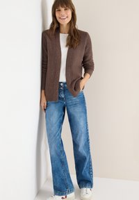 Hnědý žebrovaný svetr na knoflíky přes bílé tričko, doplněný širokými modrými džíny a bílými teniskami s červenými detaily. Ležérní outfit s kapsami.