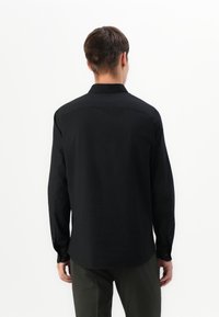 Hombre con una camiseta de manga larga negra y pantalones oscuros, de pie de espaldas contra un fondo blanco liso.