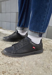 Baskets en cuir noir avec lacets plats, semelles en caoutchouc et une petite étiquette rouge Levi's. Portées avec un jean denim foncé retroussé et des chaussettes blanches montantes.