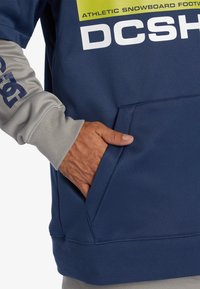 Marinblå sweatshirt med ljusgrå ärmar, med en känguruficka. Visar stor vit text och en grön logotyp på bröstet.