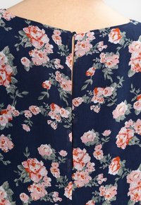 Tissu bleu marine à imprimé floral rose et rouge, fixé au centre en haut par un petit bouton noir avec une fermeture à boucle.