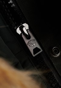 Zbliżenie na srebrny metalowy suwak zamka błyskawicznego z logo Superdry na czarnej tkaninie i czarnej taśmie zamka.