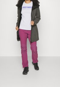 Cappotto lungo grigio con una tasca interna nera, abbinato a pantaloni rosa con toppe sulle ginocchia e scarponi da trekking neri. Maglietta bianca con logo.