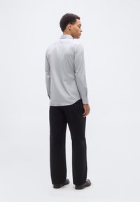 Άνδρας που στέκεται με το σώμα στραμμένο πλάγια, φορώντας ανοιχτό γκρι μακρυμάνικο πουκάμισο, μαύρο παντελόνι και σκούρα παπούτσια, σε λευκό, απλό φόντο.