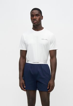 Мъж, носещ бяла тениска Calvin Klein и тъмносини къси панталони с колан на Calvin Klein, стоящ на светъл фон.