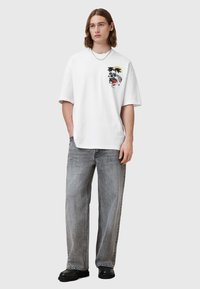 Hvit bomullstrøye med et grafisk trykk av en tiger, palmer og en sol på venstre bryst. Kombinert med lys grå, romslige jeans.