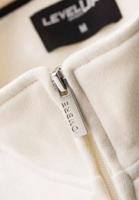 Veste zippée blanche au texture douce, dotée d'une tirette de zip en métal argenté gravée « OMBRIE » et d'un label noir indiquant « LEVEL UP », taille M.