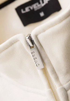Witte jack met ristsluiting en een zachte textuur, voorzien van een zilveren ritssluiting met de gravure "OMBRIE" en een zwart label met de tekst "LEVEL UP", maat M.