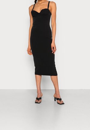 Robe noire midi avec une silhouette ajustée, des bretelles fines et des détails de bonnets texturés. Associée à des escarpins noirs à bout ouvert. Tissu lisse.