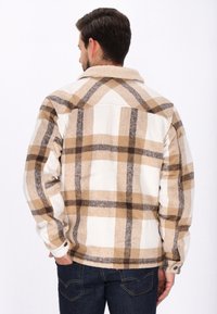 Tikkupaita, jossa on beige, ruskea ja valkoinen värimaailma, valmistettu teksturoidusta kankaasta. Siinä on sherpa-kaulus ja napit hihansuissa.