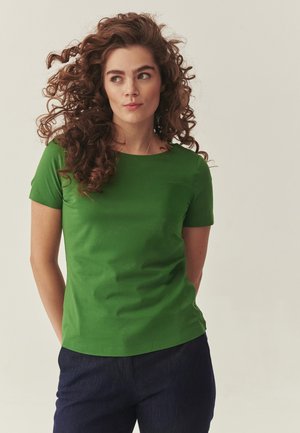 Kobieta o kręconych włosach, ubrana w zieloną koszulkę z krótkim rękawem i ciemne spodnie, spoglądająca lekko na bok na tle jednolitego tła.