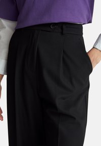 Zwarte broek met een getailleerde pasvorm, voorzien van voorplooien, zijzakken en een gladde textuur. Gestyled met een paarse blouse en witte mouwen.
