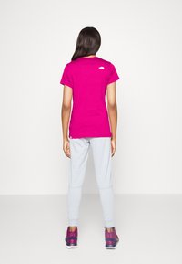 Camiseta de manga corta fucsia, pantalones deportivos grises y zapatos deportivos morados. La camiseta tiene un pequeño logo en la parte superior de la espalda.