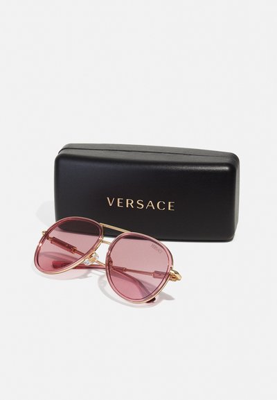 Versace saulesbrilles ar rozā tonētiem stikliem, zelta metāla rāmis un smalkiem apdariem, komplektā ar melnu futlāri ar uzrakstu "Versace."