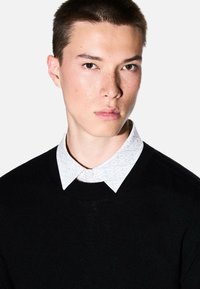 Maglione nero con scollo tondo indossato sopra una camicia bianca con colletto decorata da un motivo geometrico nero. Tessuto liscio, design moderno.