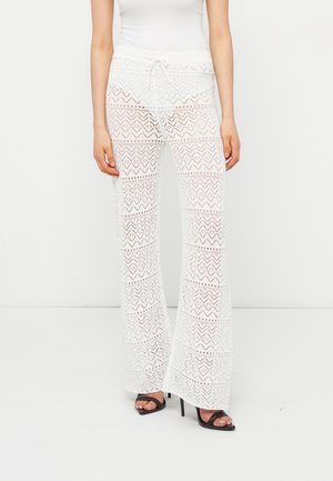 Kobieta ubrana w białe, koronkowe spodnie o szerokich nogawkach z geometrycznymi wzorami, w połączeniu z czarnymi, odkrytymi na palce butami na wysokim obcasie i białym, bezrękawnym topem.