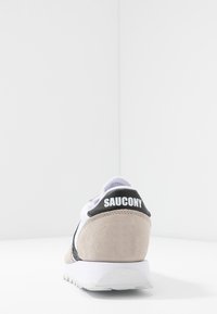 Beżowe i białe sneakersy Saucony zamszowe, z czarnym paskiem z logo na pięcie, teksturowanym panelem oraz białą gumową podeszwą.