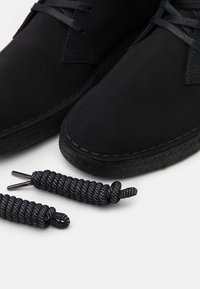 Svarta mockaskor med texturerad yta och tjocka svarta gummisulor, fyllda med matchande svarta och vita fläckiga snören.