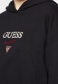 Svart hoodie med broderad "GUESS"-logotyp i vitt och "originals" i rött, med en triangel-logotypdetalj på framsidan. Mjuk tygtextur.