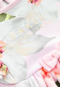 Abito con motivo floreale in tessuto rosa chiaro e bianco, caratterizzato da scritte dorate "BAKER BY TED BAKER" e un piccolo accento a forma di fiocchetto rosa.