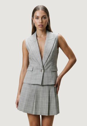 Kvinde med glat hår bærer en ærmeløs grå ternet blazer og matchende plisseret nederdel, står med den ene hånd på hoften.
