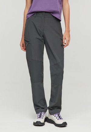 Persona che indossa pantaloni da esterno grigio scuro con tasca laterale con zip e scarpe da trekking bianche con lacci viola.