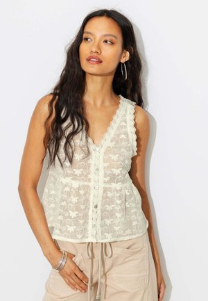 Femme aux longs cheveux foncés portant un top sans manches en dentelle blanche à boutons et un pantalon beige avec des poches, regardant sur le côté.
