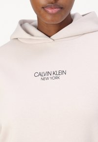 Sudadera de color beige claro, fabricada con un material suave, que presenta un logo en el frente que dice "CALVIN KLEIN NEW YORK" en letras oscuras.