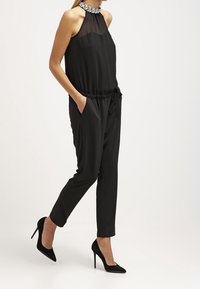 Mono negro con cuello halter, tejido transparente en la parte superior y un escote decorativo. Pantalones ajustados con bolsillos laterales, combinados con tacones negros.