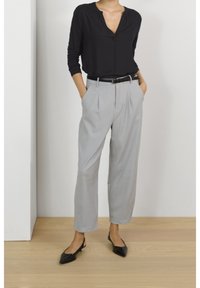 Kvinde står med hænderne i lommerne iført en sort langærmet top, lys grå plisserede bukser, sort bælte og sorte spidse sko.