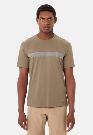 Uomo che indossa una maglietta a maniche corte di colore marrone chiaro con strisce orizzontali blu e beige sul petto, abbinata a pantaloni beige.