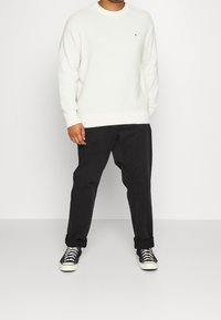 Maglione bianco lavorato a maglia con texture a coste, abbinato a pantaloni neri e scarpe da ginnastica bianche e nere. Logo piccolo e distintivo sul petto del maglione.