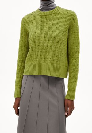 Personne portant un pull tricoté texturé vert citron sur un haut gris à col montant et une jupe plissée grise, debout les bras détendus.