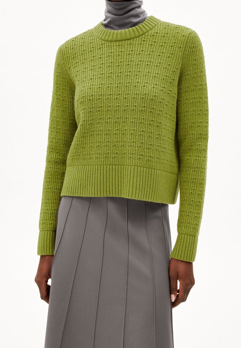 Personne portant un pull tricoté texturé vert citron sur un haut gris à col montant et une jupe plissée grise, debout les bras détendus.