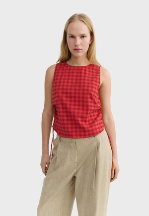 Femme portant un haut rouge à carreaux sans manches et un pantalon beige ample, debout devant un fond uni gris clair.